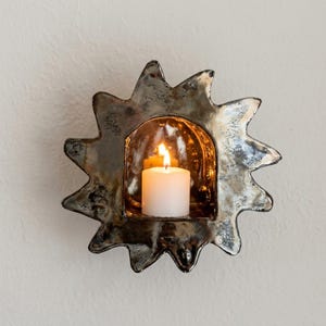 May include: A sun-shaped wall-mounted candle holder with a metallic, reflective finish. The center features an arched opening containing a small, lit white pillar candle that casts a warm glow against the interior of the dark, textured frame.