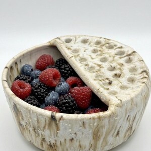 May include: A round, off-white ceramic bowl with a textured, brown-streaked glaze and a semi-circular lid featuring multiple circular holes. The open section of the bowl is filled with a mixture of fresh red raspberries, blueberries, and blackberries.