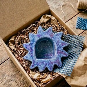 May include: A star-shaped ceramic dish with a glossy finish in shades of blue and purple. The dish features a central arched indentation and pointed rays extending outward, presented inside a cardboard box filled with brown paper filler.