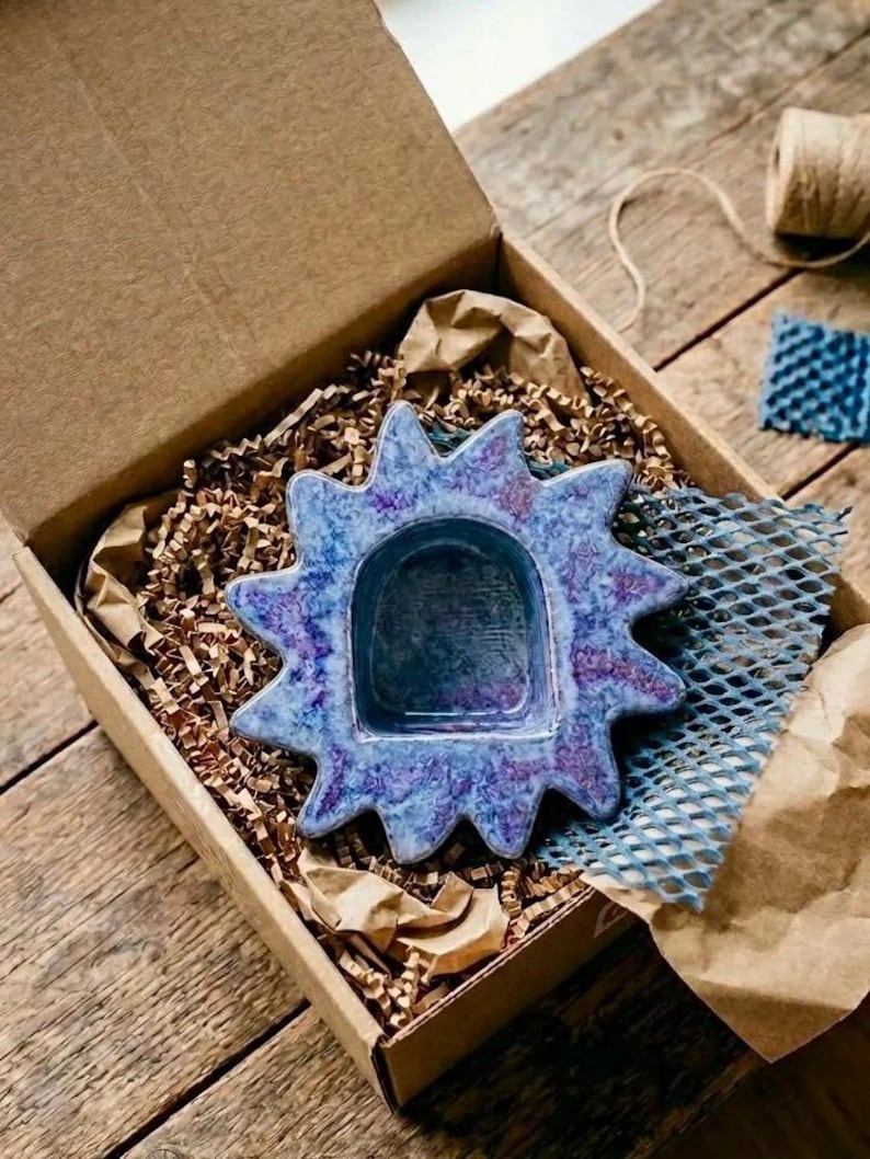 May include: A star-shaped ceramic dish with a glossy finish in shades of blue and purple. The dish features a central arched indentation and pointed rays extending outward, presented inside a cardboard box filled with brown paper filler.