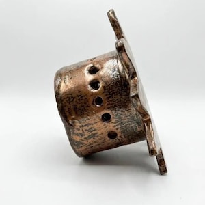 May include: A cylindrical ceramic object with a metallic bronze finish and a textured surface. The piece features a wide, flat, star-shaped rim and a series of small, circular perforations arranged in a vertical line along the side of the body.