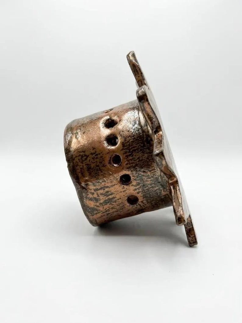 May include: A cylindrical ceramic object with a metallic bronze finish and a textured surface. The piece features a wide, flat, star-shaped rim and a series of small, circular perforations arranged in a vertical line along the side of the body.