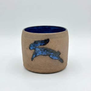May include: A cylindrical ceramic cup with a textured, matte tan exterior and a smooth, deep blue interior. The outer surface features a dark blue, hand-painted silhouette of a leaping rabbit, contrasting with the earthy, neutral clay tone.