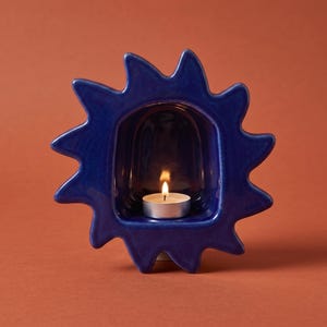 May include: A deep blue ceramic candle holder shaped like a stylized sun with a central arched opening. A small, round white tea light candle sits inside the hollow center, featuring a lit flame that casts a warm glow against the glossy blue surface.