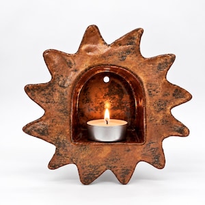 May include: A sun-shaped wall sconce crafted from textured, metallic bronze-toned ceramic. The piece features a central arched alcove containing a small, lit white tea light candle. A circular hole is positioned at the top of the inner arch.