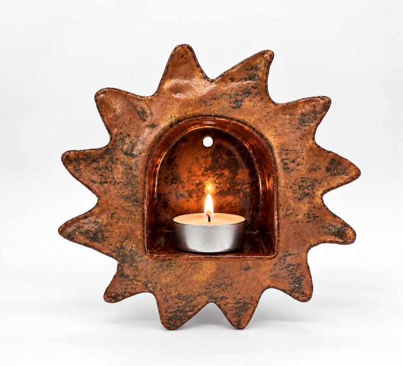 May include: A sun-shaped wall sconce crafted from textured, metallic bronze-toned ceramic. The piece features a central arched alcove containing a small, lit white tea light candle. A circular hole is positioned at the top of the inner arch.