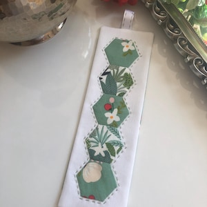 May include: A white fabric bookmark with a green, white, and red floral pattern. The bookmark is made of six hexagonal fabric pieces sewn together.