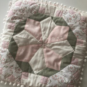 May include: Square quilted cushion with a starburst design in pink, white, and green. It features floral and gingham patterns, with a border of white pom-poms. A decorative accent for a bed or sofa, measuring approximately 40cm x 40cm.