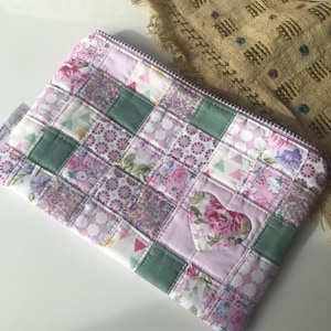 May include: A quilted patchwork zippered pouch in shades of pink, lavender, and green. The design features a heart-shaped appliqué with floral fabric. The pouch is rectangular and has a purple zipper.