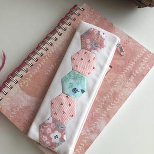 May include: White zippered pencil case with a patchwork design of pink, blue and white hexagonal fabric pieces with floral and polka dot patterns.