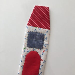 May include: A fabric bookmark in the shape of a house. The roof is red with white polka dots, the body is white with a floral pattern, and it has a blue denim window and a red door.