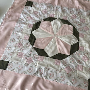 Könnte beinhalten: Eine handgefertigte Steppdecke mit einem zentralen Sternmuster in Rosa und Weiß, umgeben von einem dunkelgrünen Achteck. Die Steppdecke hat einen Stoff mit Blumenmuster und einen rosafarbenen Satinrand mit weißer Bommelborte. Die Steppdecke ist quadratisch.
