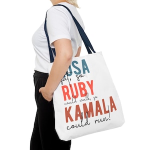 May include: A white tote bag with a navy blue handle. The bag has a graphic design with the text "USA sat, so RUBY could walk, so KAMALA could run!"