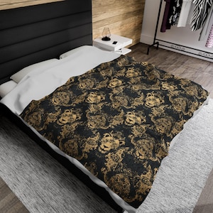 Op de afbeelding: Een zwart en gouden deken met een herhalend patroon van leeuwenkoppen. Het deken ligt op een bed met witte lakens en een zwart hoofdeinde.
