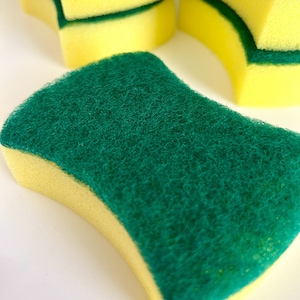 May include: Close-up of several yellow and green kitchen sponges. The sponges have a yellow base with a green, textured scrubbing surface. Some sponges are stacked, while others are in the foreground, showing their shape and texture.