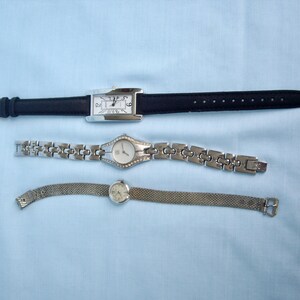May include: Three wristwatches on a light blue surface. The top watch has a rectangular silver face and a black leather band. The middle watch has a silver band with a round face. The bottom watch has a mesh band and a round face.