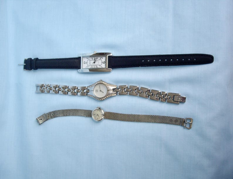 May include: Three wristwatches on a light blue surface. The top watch has a rectangular silver face and a black leather band. The middle watch has a silver band with a round face. The bottom watch has a mesh band and a round face.