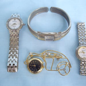 May include: An assortment of jewelry and timepieces, including two silver-toned watches with white faces, a silver and gold-toned bracelet, and a gold-toned car-shaped clock with a black face. The items are arranged on a light blue surface.
