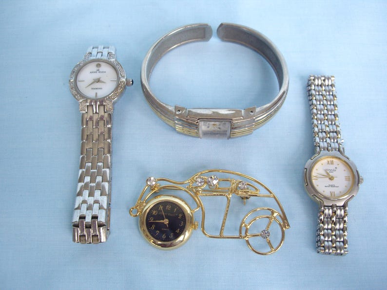 May include: An assortment of jewelry and timepieces, including two silver-toned watches with white faces, a silver and gold-toned bracelet, and a gold-toned car-shaped clock with a black face. The items are arranged on a light blue surface.