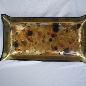 Puede incluir: Bandeja decorativa rectangular con un diseño dorado y negro. La bandeja presenta un patrón moteado en tonos marrón, dorado y negro. Los bordes están adornados con un borde dorado y un ribete negro.