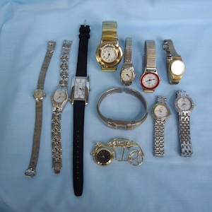 May include: A collection of various wristwatches, including gold-toned, silver-toned, and black-banded styles. Some watches have metal bands, while others have leather or fabric straps. The watches are arranged on a light blue surface.