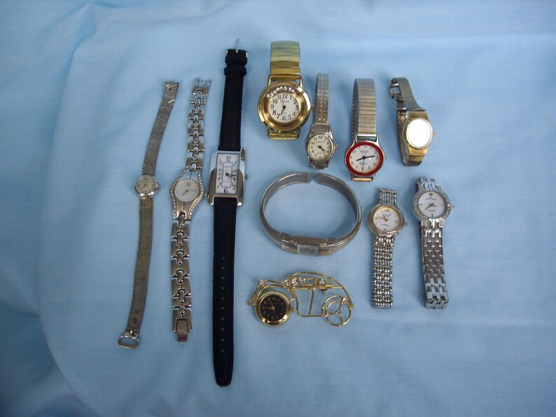 May include: A collection of various wristwatches, including gold-toned, silver-toned, and black-banded styles. Some watches have metal bands, while others have leather or fabric straps. The watches are arranged on a light blue surface.