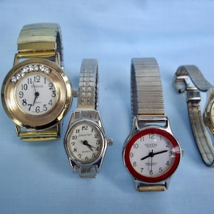May include: Four wristwatches with different designs. One has a gold-tone case and band with a white face and crystal accents. Another has a silver-tone case and band with a white face. The third has a red bezel and a white face. The last one has a gold-tone case and band.