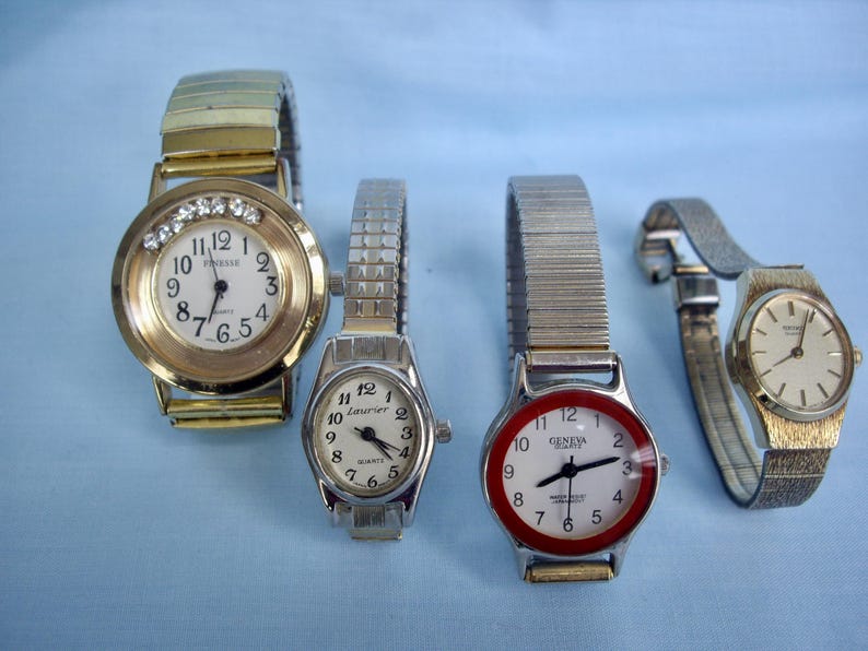 May include: Four wristwatches with different designs. One has a gold-tone case and band with a white face and crystal accents. Another has a silver-tone case and band with a white face. The third has a red bezel and a white face. The last one has a gold-tone case and band.