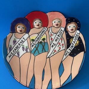 May include: A ceramic plate with a blue background features four figures in swimsuits and hats. Each figure wears a sash labeled "Ms. Cabernet", "Ms. Chardonnay", "Ms. Merlot", and "Ms. Zinfandel". The plate has a whimsical, cartoonish style.