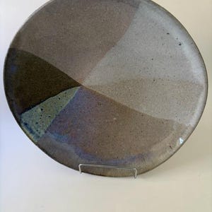 May include: A round ceramic plate featuring a speckled, multi-toned glaze with geometric color blocking in shades of brown, tan, grey, and blue. The surface has a textured, earthy appearance with a smooth, slightly curved rim and a flat center.