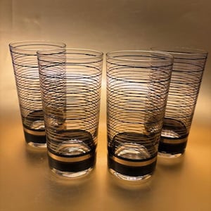 May include: Four clear highball glasses with black and gold bands and a black swirl design. The glasses have a cylindrical shape and are arranged in a group. The glasses are made of glass and have a retro design.