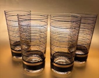 Puede incluir: Cuatro vasos altos transparentes con bandas negras y doradas y un diseño de remolino negro. Los vasos tienen forma cilíndrica y están dispuestos en grupo. Los vasos son de vidrio y tienen un diseño retro.