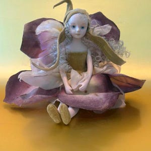 May include: A porcelain doll with pale skin and curly white hair, wearing a pointed olive-green hat and a matching bodice. The doll is seated amidst large, layered fabric petals in shades of deep purple and cream, resembling a flower blossom.