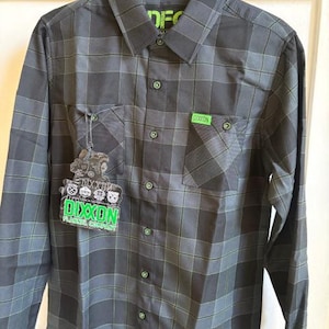May include: A long-sleeved, button-down flannel shirt in a gray and black plaid pattern with green accents. The shirt has two chest pockets and a small green label that says "DIXXON". A hang tag with the brand name is attached.