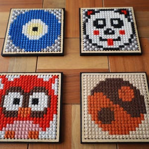 May include: Four square coasters with cross-stitch designs. One features a blue eye, another a panda, an owl, and a mushroom. Each coaster has a black border and is set on a wooden surface.