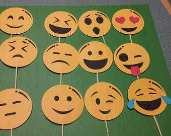 May include: A set of 12 yellow paper emoji face cutouts with different expressions, including happy, sad, surprised, and winking. The cutouts are attached to wooden sticks.