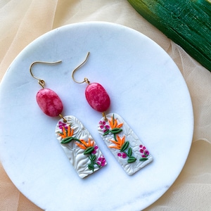 May include: A pair of dangle earrings with pink and white clay flowers and a pink stone charm. The earrings have a gold hook.