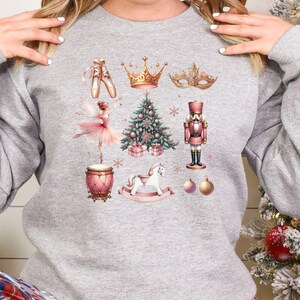 May include: A light gray sweatshirt featuring watercolor illustrations of ballet shoes, a crown, a masquerade mask, a ballerina, a Christmas tree, a nutcracker, a drum, a rocking horse, and ornaments. The design has a festive, holiday theme.