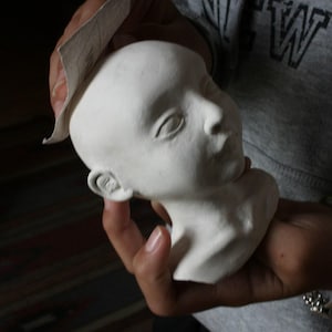 May include: A white, unpainted ceramic head of a person, held in a person's hands. The head is smooth and features detailed facial features, including eyes, a nose, and a mouth. A piece of sandpaper is held near the head.