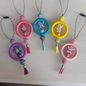 May include: Six Care Bear-themed accessories in various colours. Each has a Care Bear figure inside a ring, a beaded accent, and a tassel. The rings are pink, blue, yellow, purple, and light pink. The accessories hang from black cords.