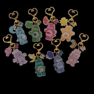 May include: A selection of Care Bear keyrings in various colours, each with a gold-coloured heart-shaped clasp. Each keyring showcases a different Care Bear character with unique designs and accessories, including rainbows, hearts, and butterflies. The background is black.