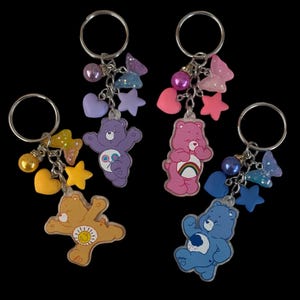May include: Four Care Bear keyrings with silver rings. Each keyring features a different coloured Care Bear charm: tan, purple, pink, and blue. Each keyring has a cluster of charms including hearts, stars, and beads in coordinating colours.
