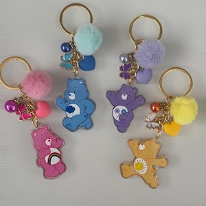 May include: Four keychains featuring cartoon bear charms in pink, blue, purple, and yellow. Each keychain includes a matching fuzzy pom-pom, a heart-shaped charm, a butterfly charm, a round bead, and a gold-toned metal ring for attachment.