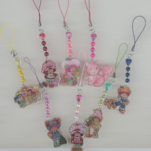 May include: A collection of eight acrylic charms featuring various cartoon characters, each attached to a colourful cord loop. The charms are decorated with small beads in matching colours, including pink, yellow, green, and blue, and depict figures with hats and pets.
