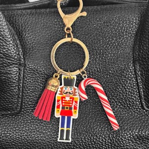 May include: A gold-toned metal keyring featuring a lobster clasp and a circular ring. Attached are three charms: a red fabric tassel with a gold cap, a flat nutcracker figure in red, blue, and gold, and a red and white striped candy cane charm.