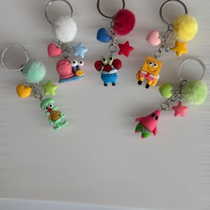 May include: A selection of keyrings featuring cartoon characters. Each keyring has a silver ring, a fluffy pom-pom, and charms such as hearts and stars. The characters include a snail, a crab, SpongeBob, Squidward, and Patrick.