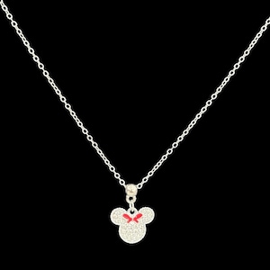 May include: Silver necklace with a mouse head pendant and a red bow. The pendant is covered in small crystals. The chain is delicate and hangs against a black backdrop. The necklace is a piece of jewellery.