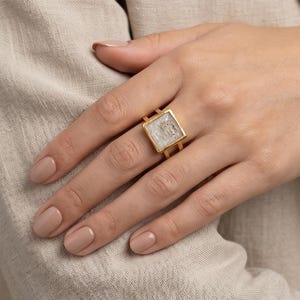 Può includere: Un anello color oro caratterizzato da una montatura quadrata con un inserto bianco traslucido e testurizzato. La fascia dell'anello è divisa in due sottili linee parallele che si collegano alla cornice quadrata, creando un design geometrico aperto.