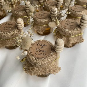 May include: Small glass jars filled with honey, each topped with a burlap lid, wooden honey dipper, and dried floral accents. The jars have a round label with the text "Cansu & Ozcan 11.02.23".