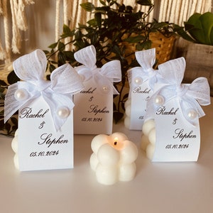 May include: Four white favour candles with a bubble design. Each candle has a white satin bow and a white card with the text "Rachel & Stephen 05.10.2024".